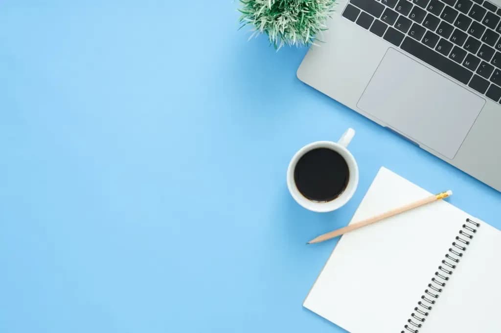 Computadora portátil, taza de café y cuaderno de espiral con lápiz sobre un escritorio azul