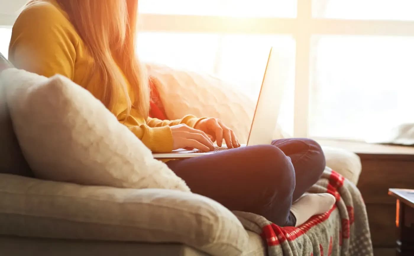 Woman with red hair working on laptop on couch by sunny window, wearing yellow cardigan