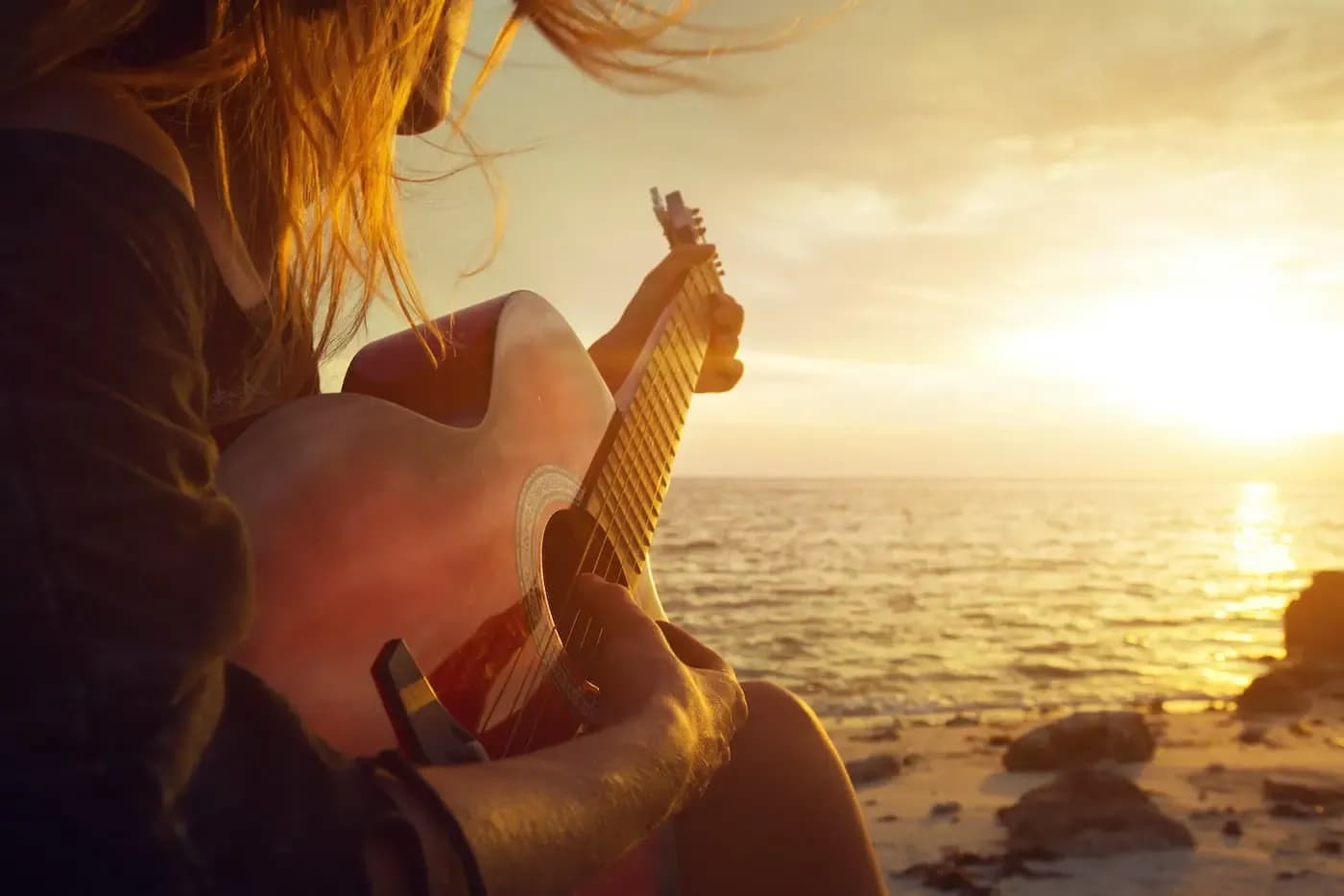 Persona tocando una guitarra acústica en la playa durante un atardecer dorado sobre el océano