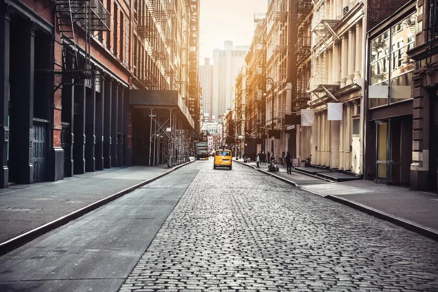Calle soleada de adoquines en la ciudad de Nueva York, con un taxi amarillo y edificios históricos a ambos lados