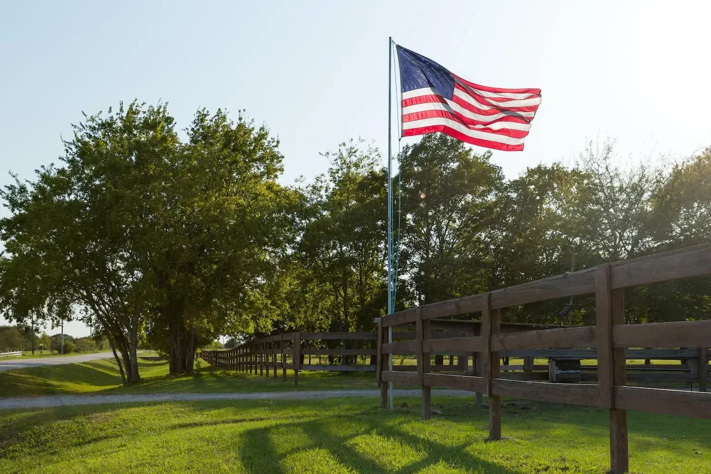 American flag waving on pole with wooden fence and trees in rural landscape