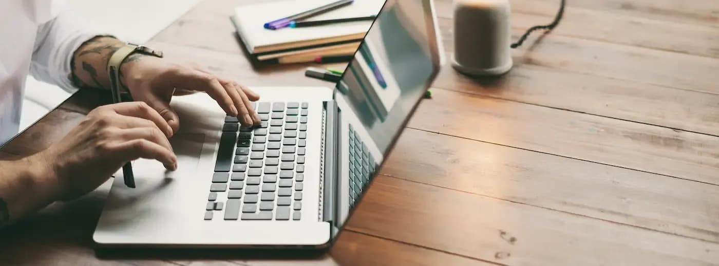 Persona escribiendo en una computadora portátil sobre un escritorio de madera con cuadernos y artículos de oficina
