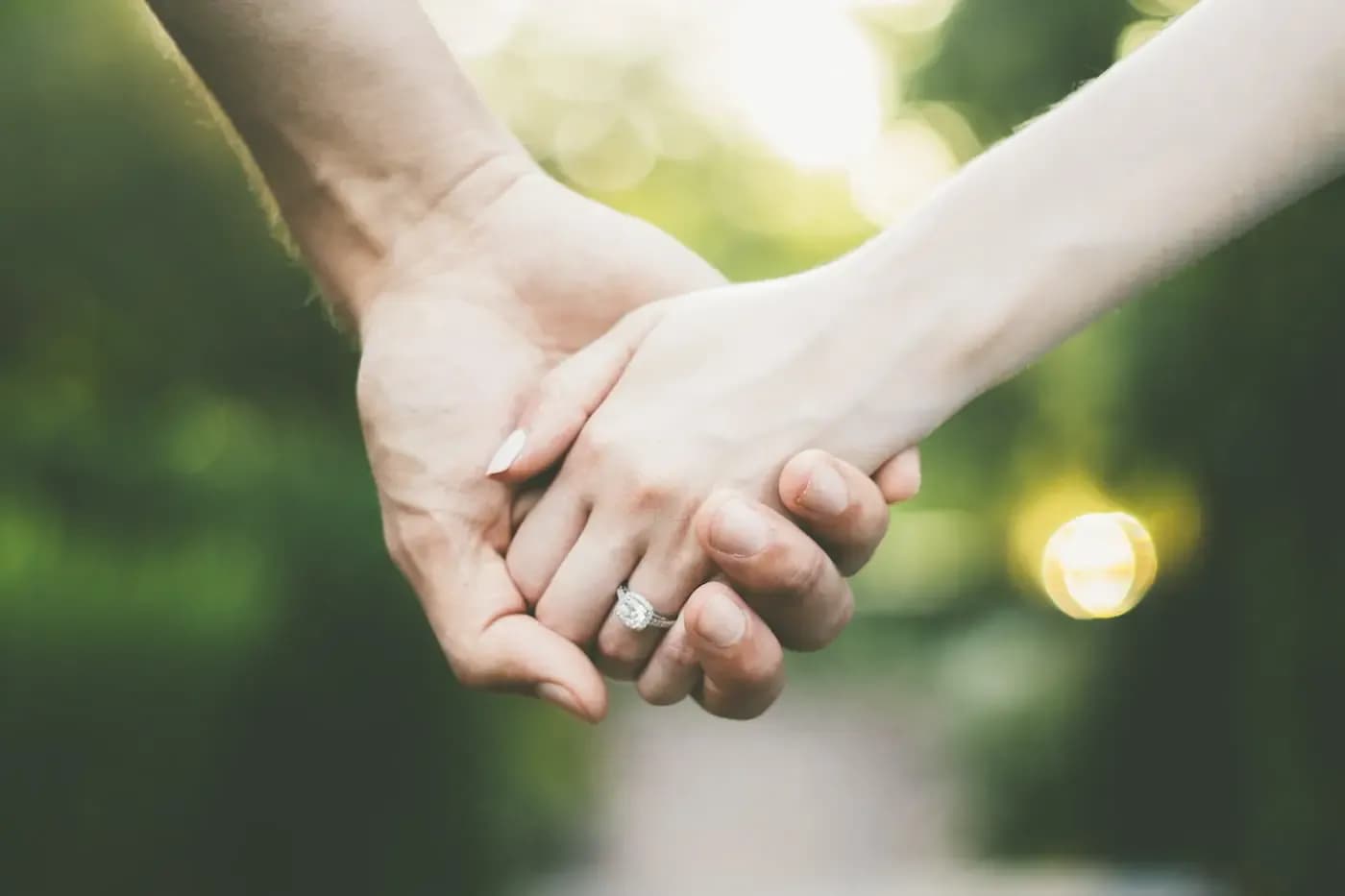 Close-up of two hands holding together outdoors, woman wearing engagement ring with blurred green background