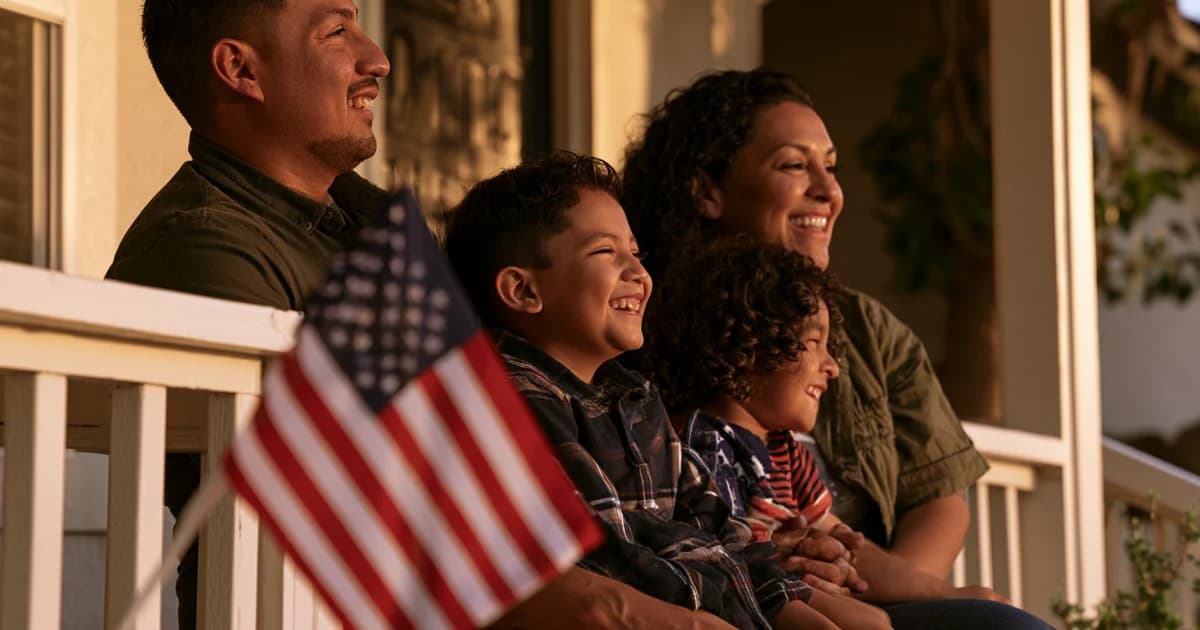 Family of four smiling together on porch with American flag at golden hour