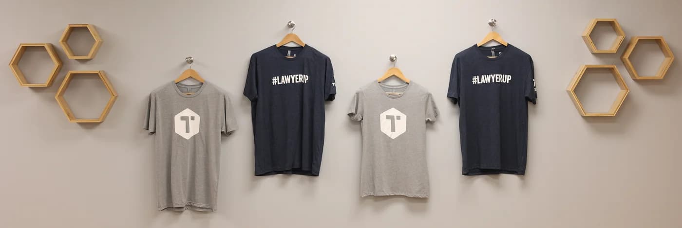 Four t-shirts on hangers displaying law firm branding and #LAWYERUP hashtag with hexagonal wall decor