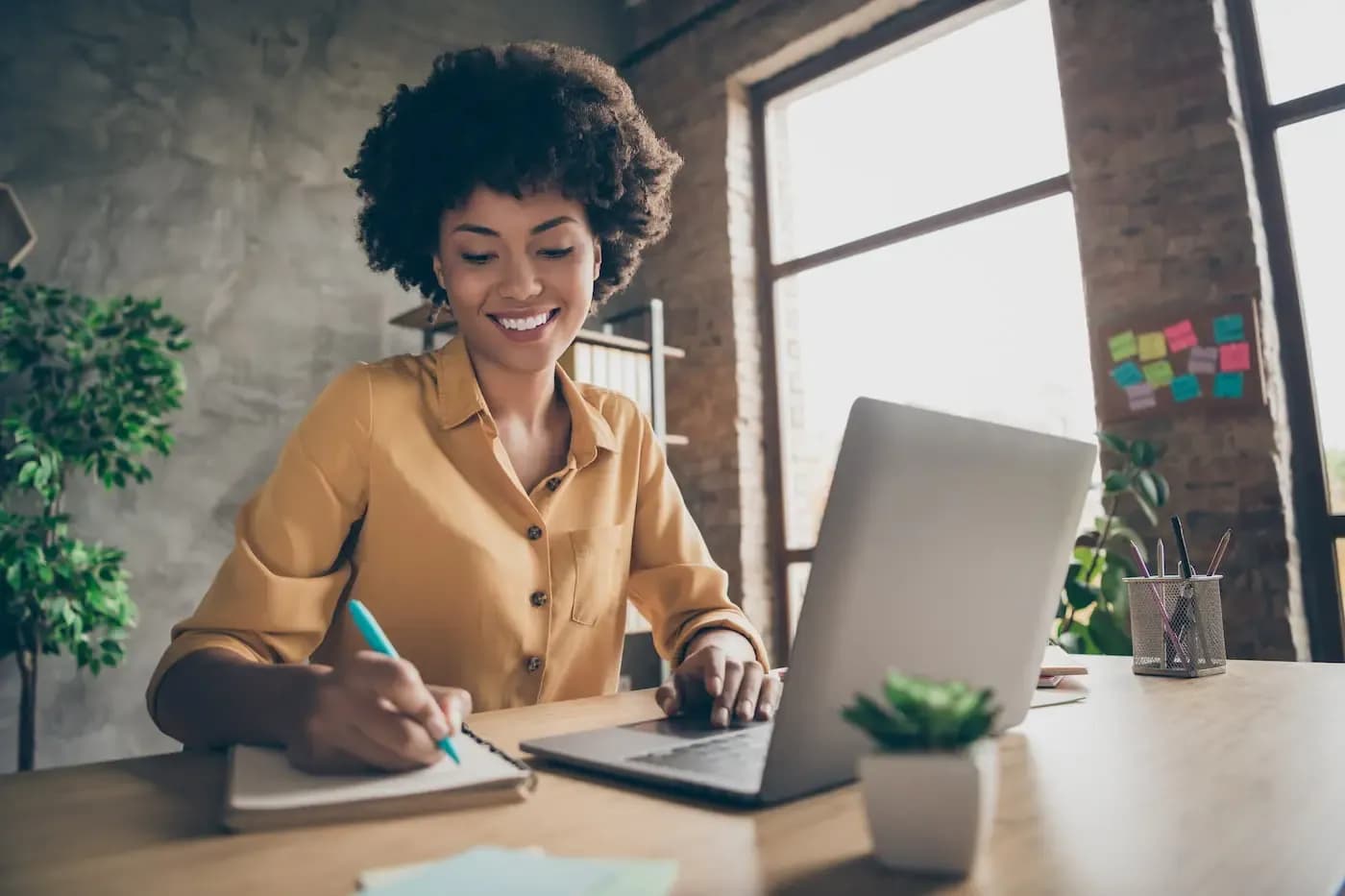 Mujer sonriente con camisa amarilla trabajando en una computadora portátil, en un escritorio de una oficina luminosa con plantas