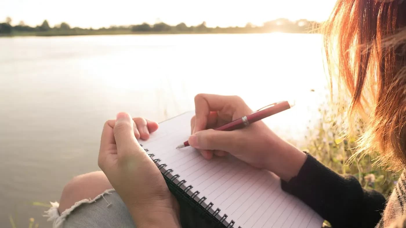 Person with reddish hair writing in a notebook by a calm lake at sunset, with trees visible across the water