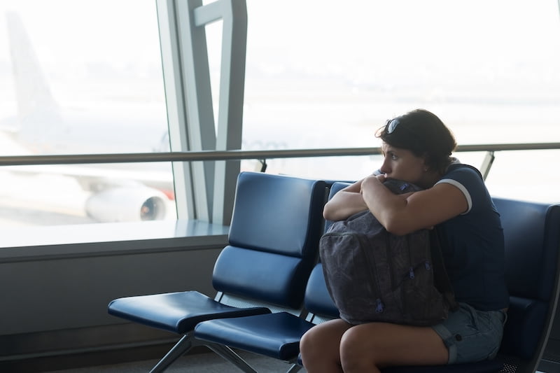 Tired caucasian woman in airport hall. Girl waiting her flight at airport terminal, holding her bag in hands, sitting on chair and looking on the window.